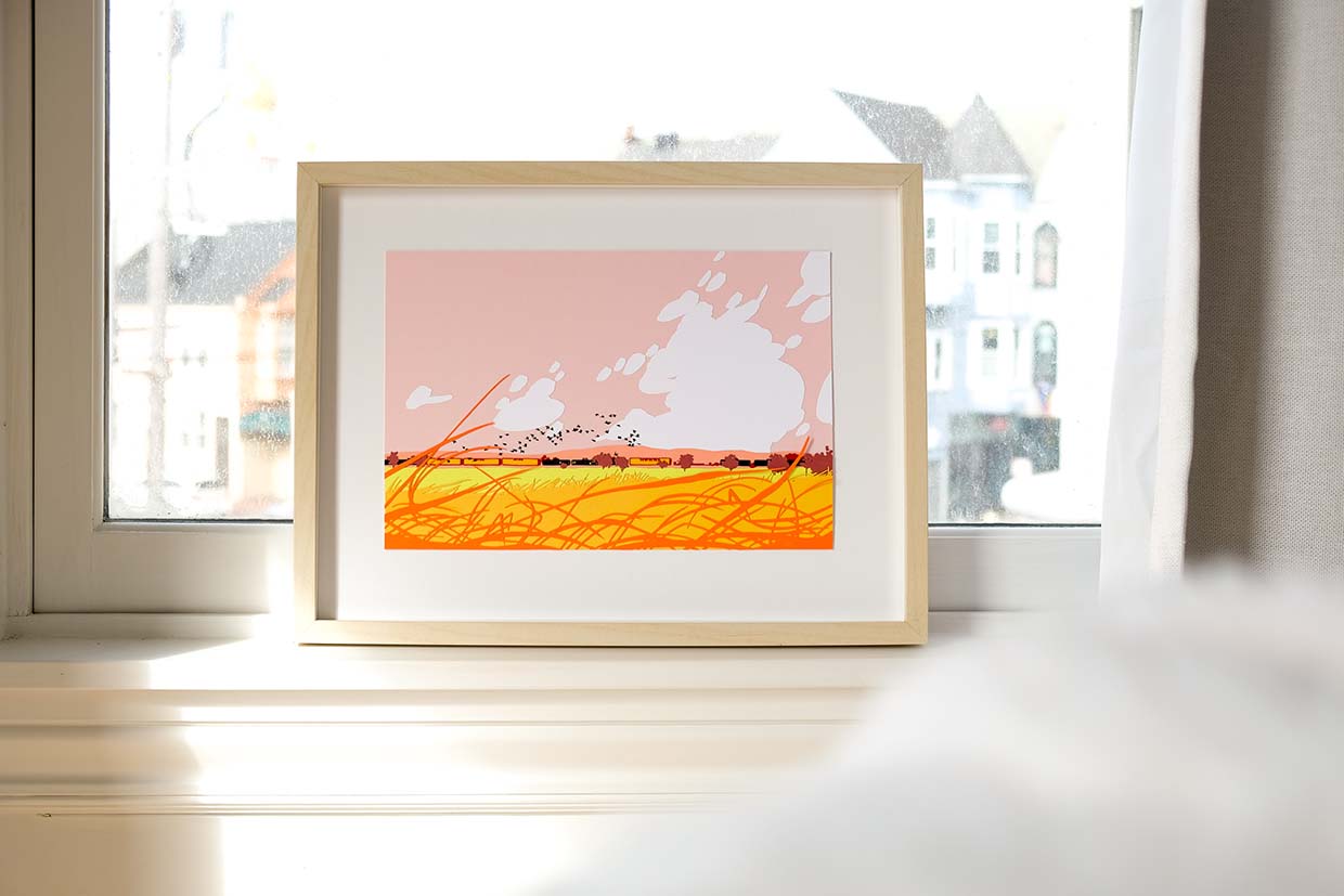 A paper train runs through the golden grasses of the Sacramento Valley under a warm pink sky. It sits in a wooden frame on a window, with a blurred view of buildings behind it.