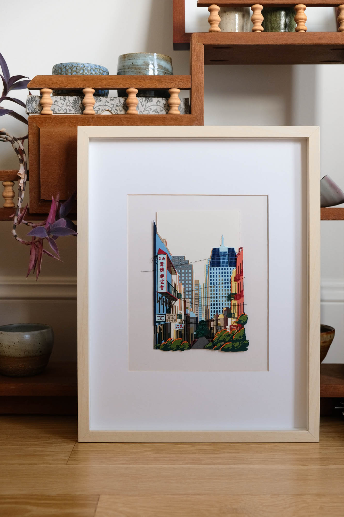 A street at the border of Chinatown and the Financial District is recreated in layers of colored papers and thread. The artwork is framed in light wood, and it sits on a hardwood floor against a vintage stepped shelf.