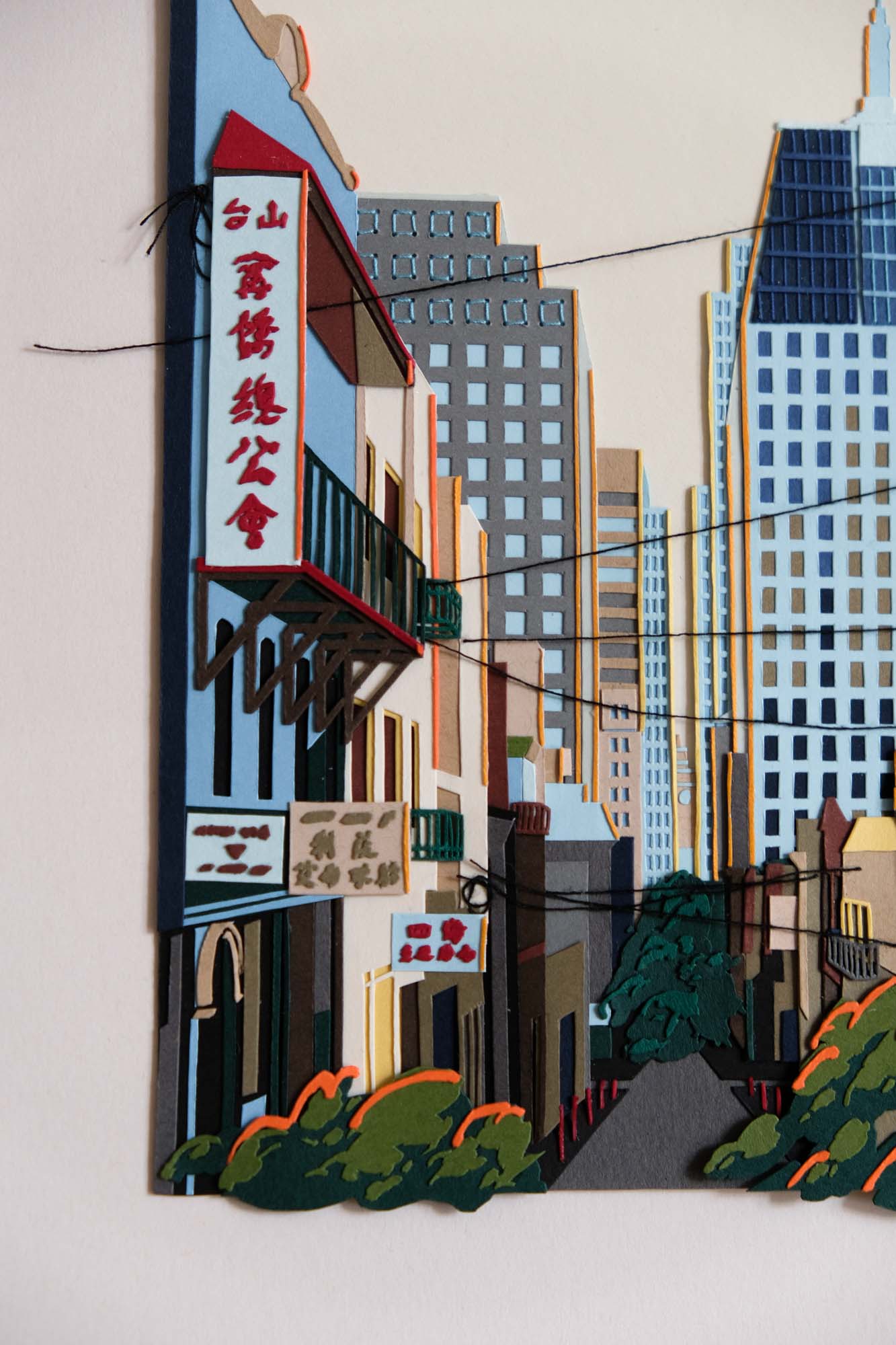 A detail view of the artwork shows black threads crisscrossing in front of paper trees and buildings. Some buildings have signs with Chinese characters.