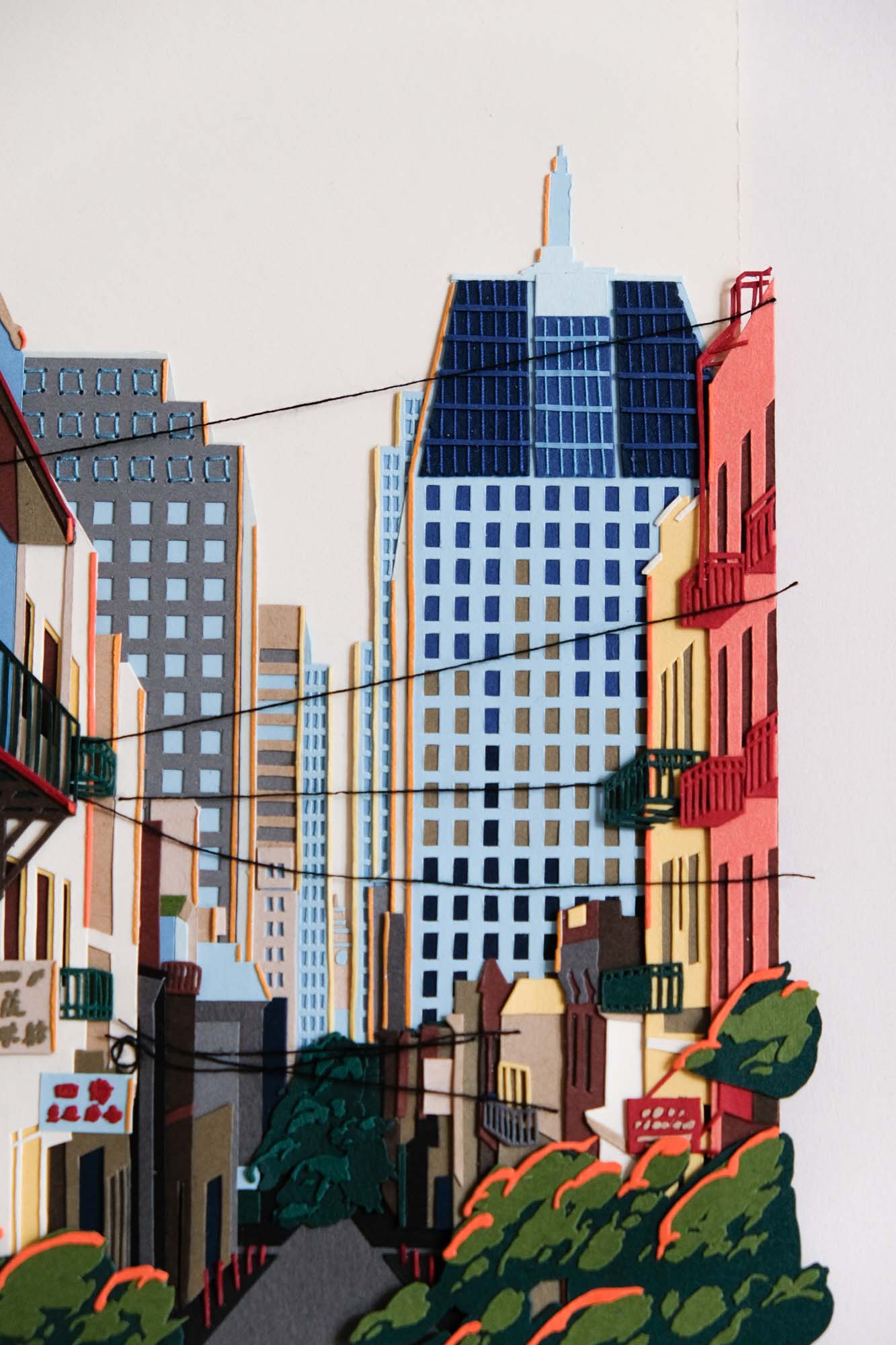 A detail view of the artwork shows black threads crisscrossing in front of paper trees and buildings. Some buildings have delicate balconies, while skyscrapers are covered in tiny windows.