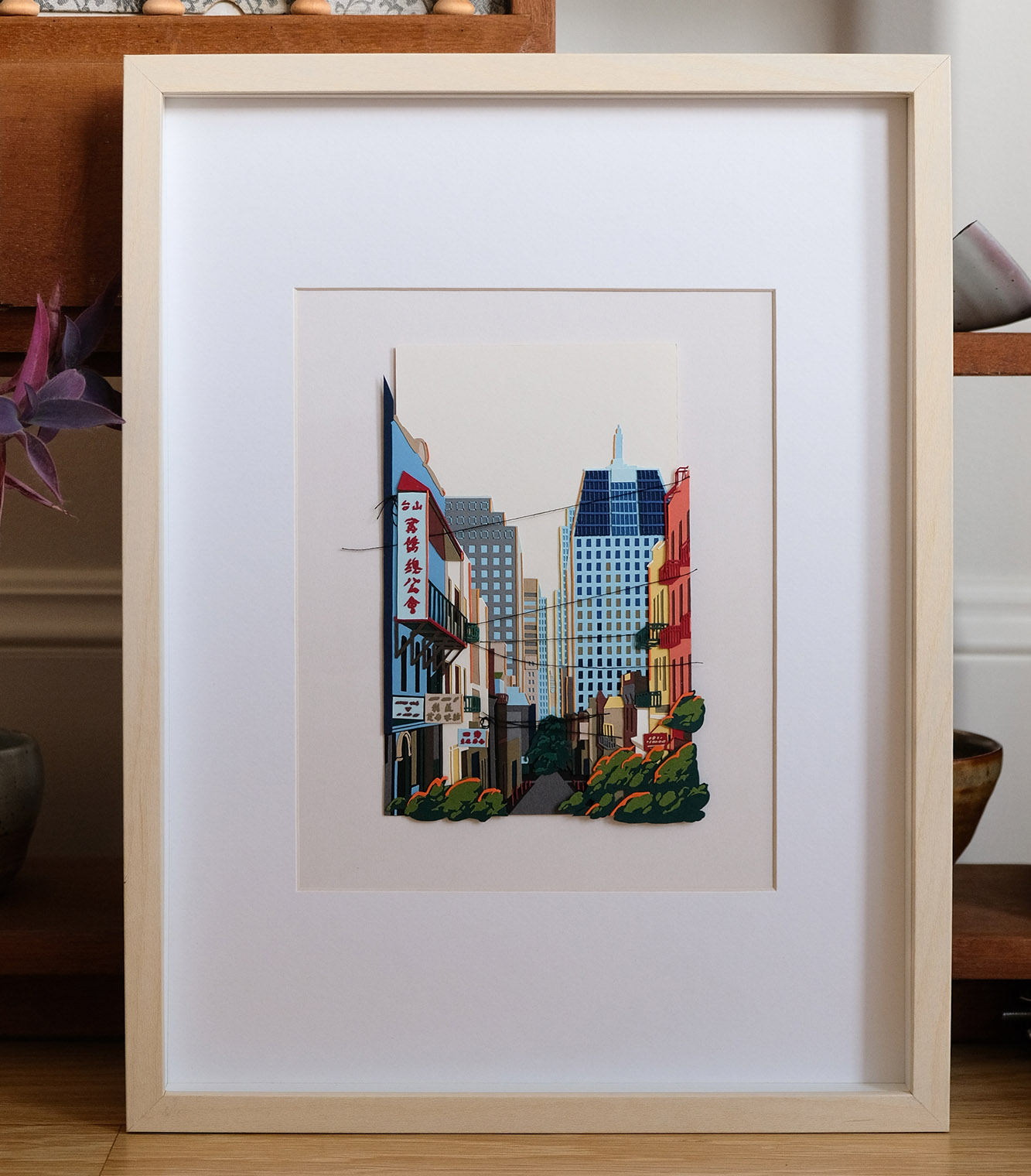 A street at the border of Chinatown and the Financial District is recreated in layers of colored papers and thread. The artwork is framed in light wood, and it sits on a hardwood floor against a vintage stepped shelf.