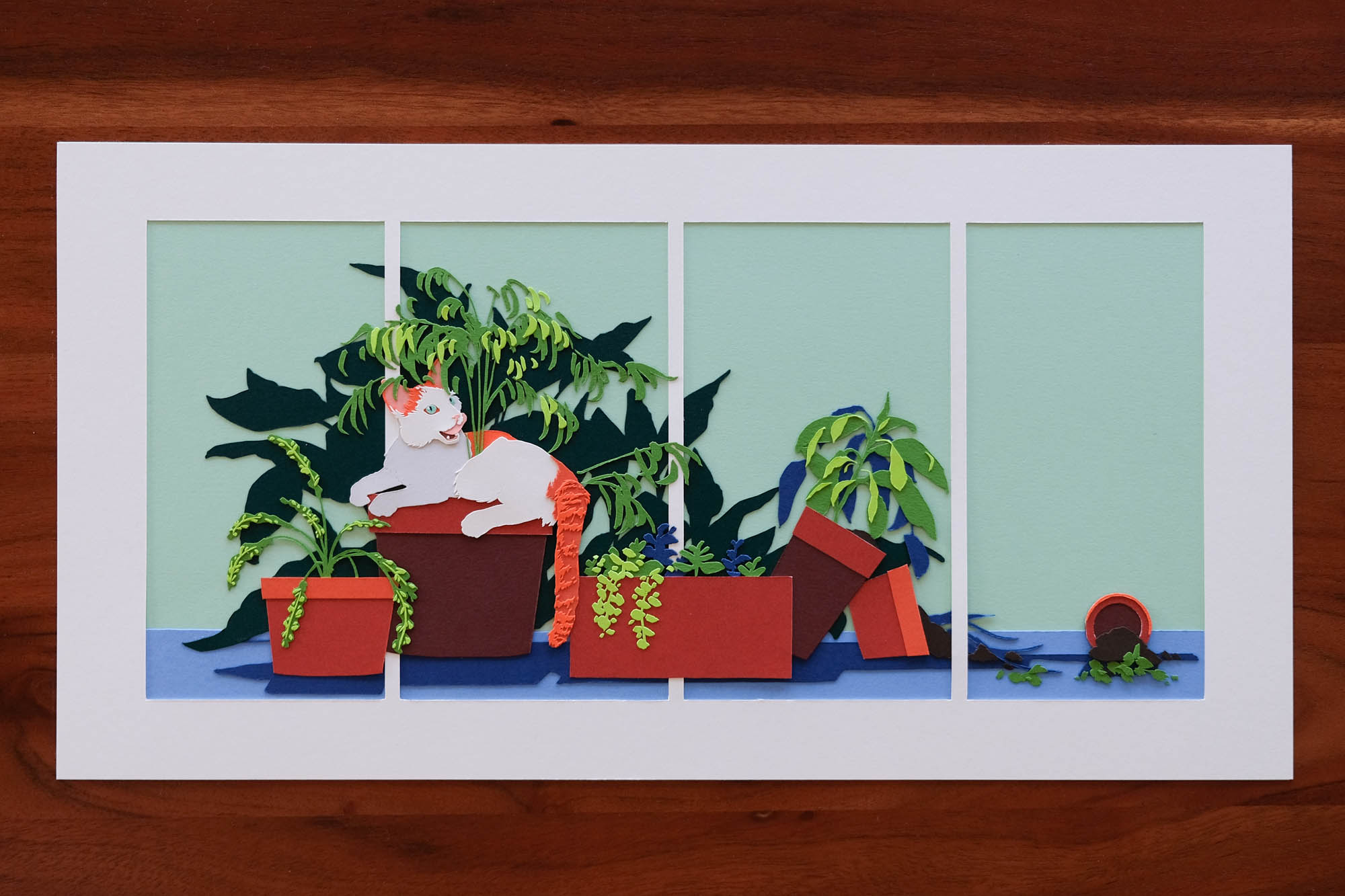 A panoramic paper portrait depicts a young orange cat reclining on a pot surrounded by paper houseplants. The pot to the far right has been knocked over. The artwork is split into four vertical sections by a custom white mat.