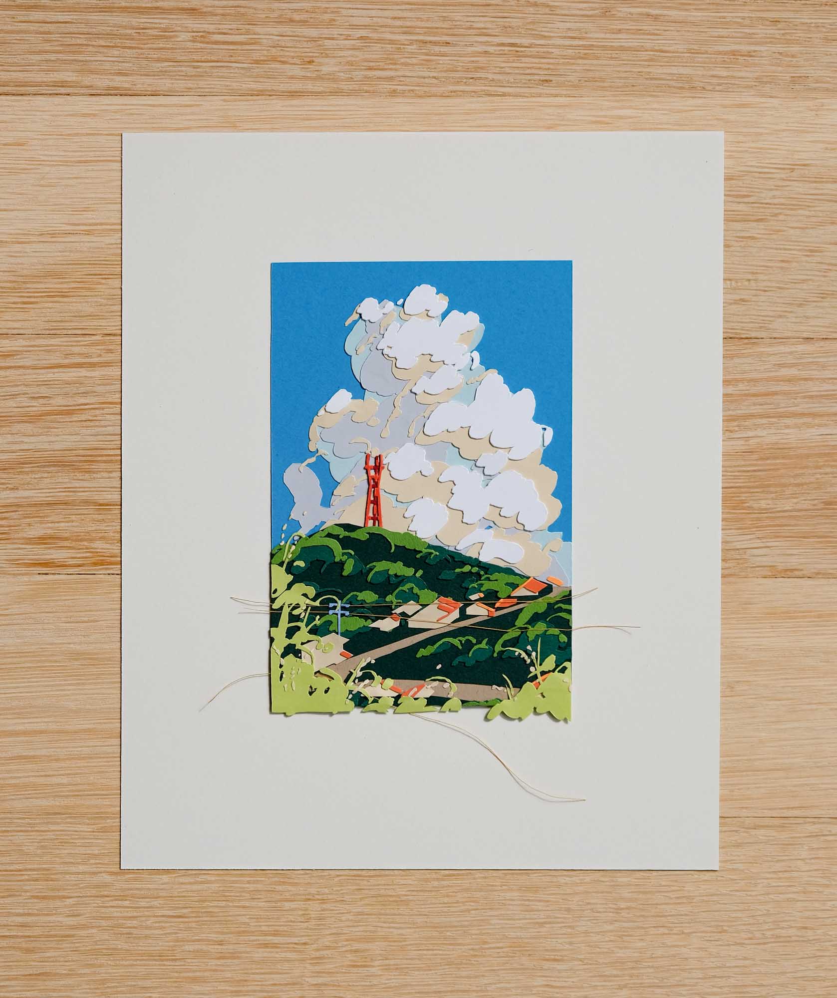Sutro Tower rises from the lush green peak of Mount Sutro in front of a large cumulus cloud and a bright blue sky. The paper artwork is framed by a large white border on a light wood surface.