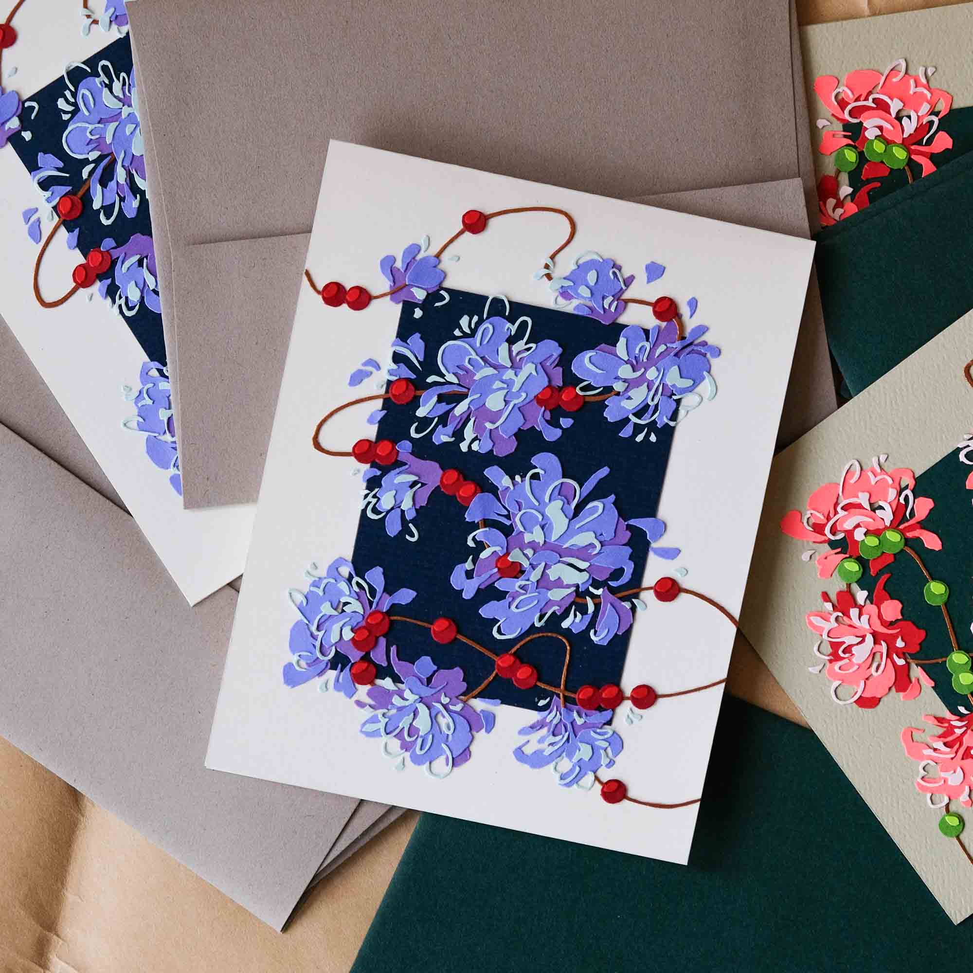 Two sets of handmade cards are decorated with expressive garlands of flowers and berries. One set has purple blooms and red accents while the other has pink blooms and light green accents. The cards are scattered among warm gray and forest green envelopes.
