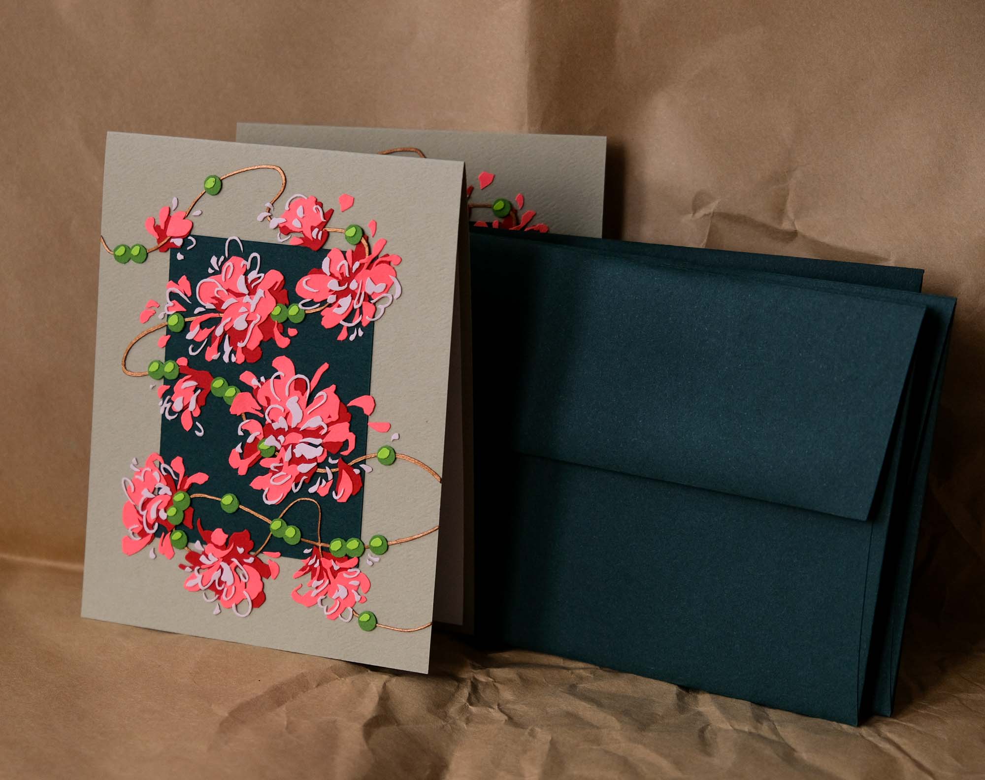 A handmade card is decorated with expressive paper garlands of pink flowers and green berries. Behind it sits a stack of dark green envelopes and a warm brown paper backdrop.