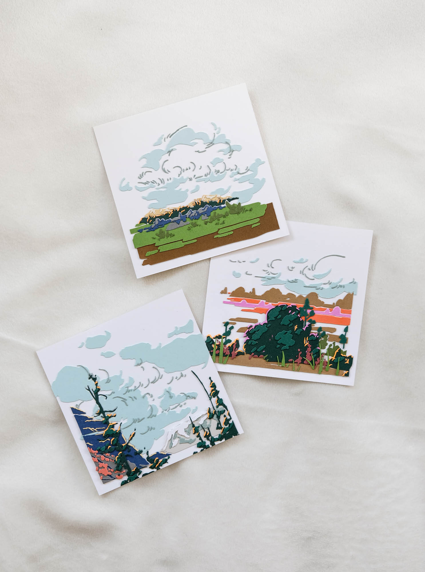 Three square artworks sit against a white backdrop. One depicts a grassy plain, the second features bushes in front of a warm landscape, and the last features treetops before a mountainous landscape. All three artworks have a prominent display of cloud cover.