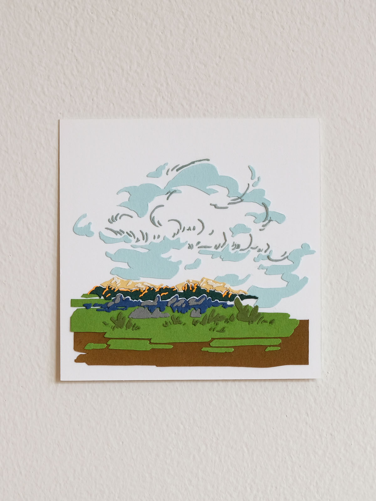 Abstracted clouds made of paper shadows and lines float across a mostly flat grass and dirt landscape, with rocks and distant mountains at the horizon. The square artwork floats on a white wall.