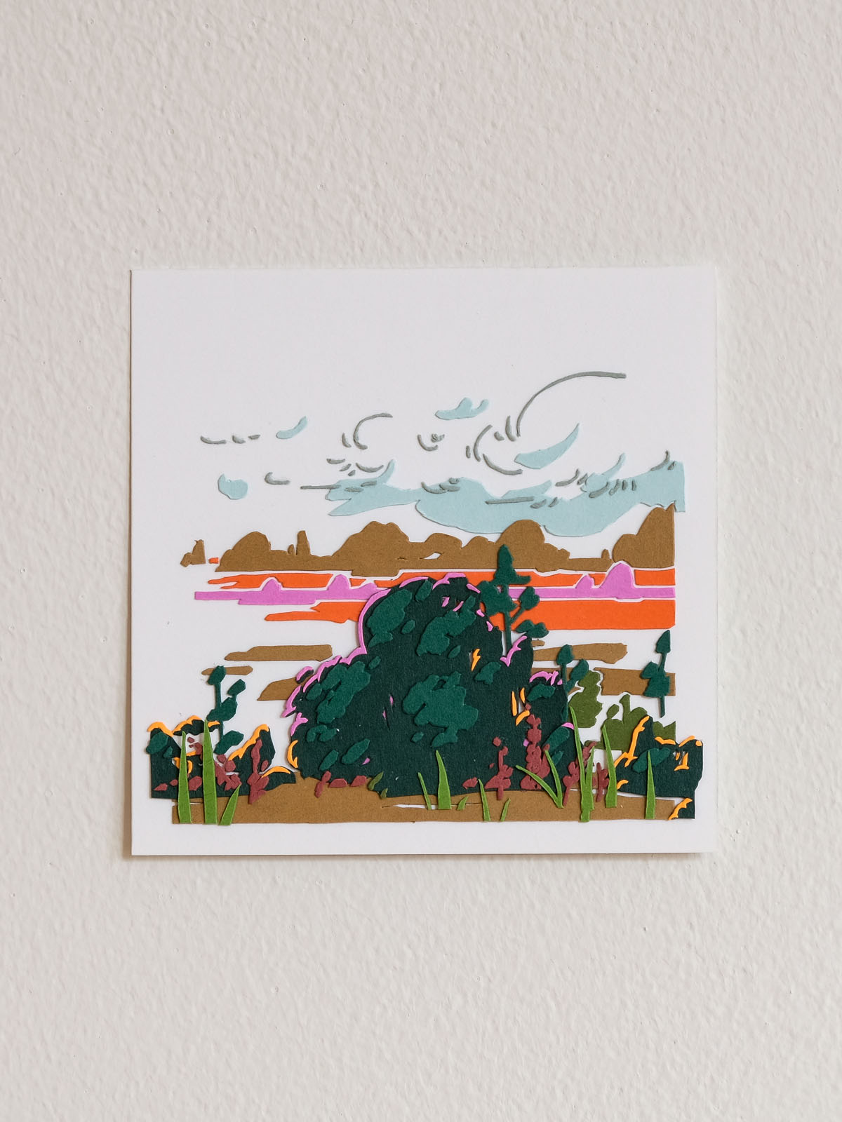 Abstracted clouds made of paper shadows and lines glide above large clusters of fauna at the edge of a warm-colored landscape. The square artwork floats on a white wall.