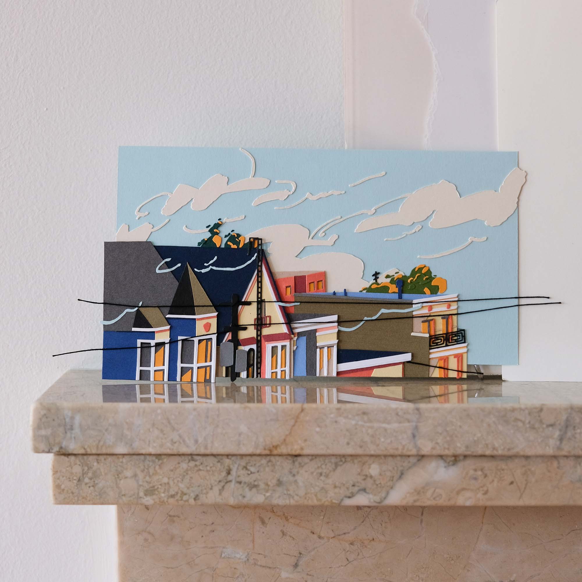 A row of paper buildings in blues, browns, and whites reflect early sunset tints of yellows, oranges, and pinks in front of a light blue paper sky streaked with pale clouds. The artwork rests on a polished stone mantle against a white wall.