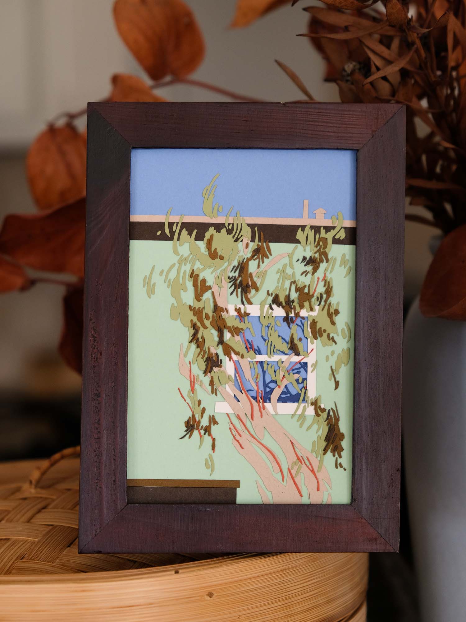 In front of a mint green suburban house, a crooked tree with gestural leaves covers a blue window. The paper artwork sits in a dark wood frame next to dried leaves and bamboo décor.
