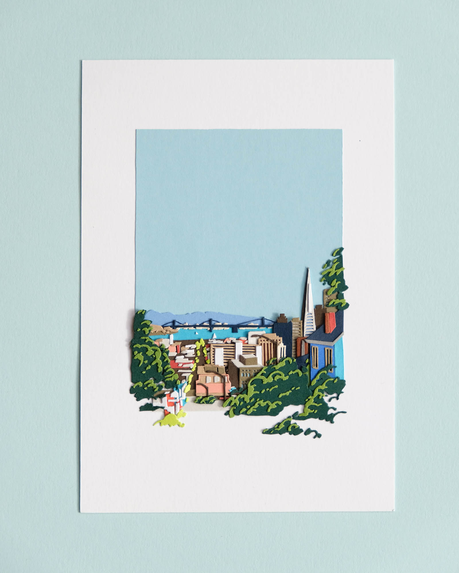 Paper trees and foliage frame distant buildings, apartments, and skyscrapers in front of an even more distant Bay Bridge and the blue expanse of the San Francisco Bay. The composition is framed on a white sheet of paper and sits on top of a light blue background.