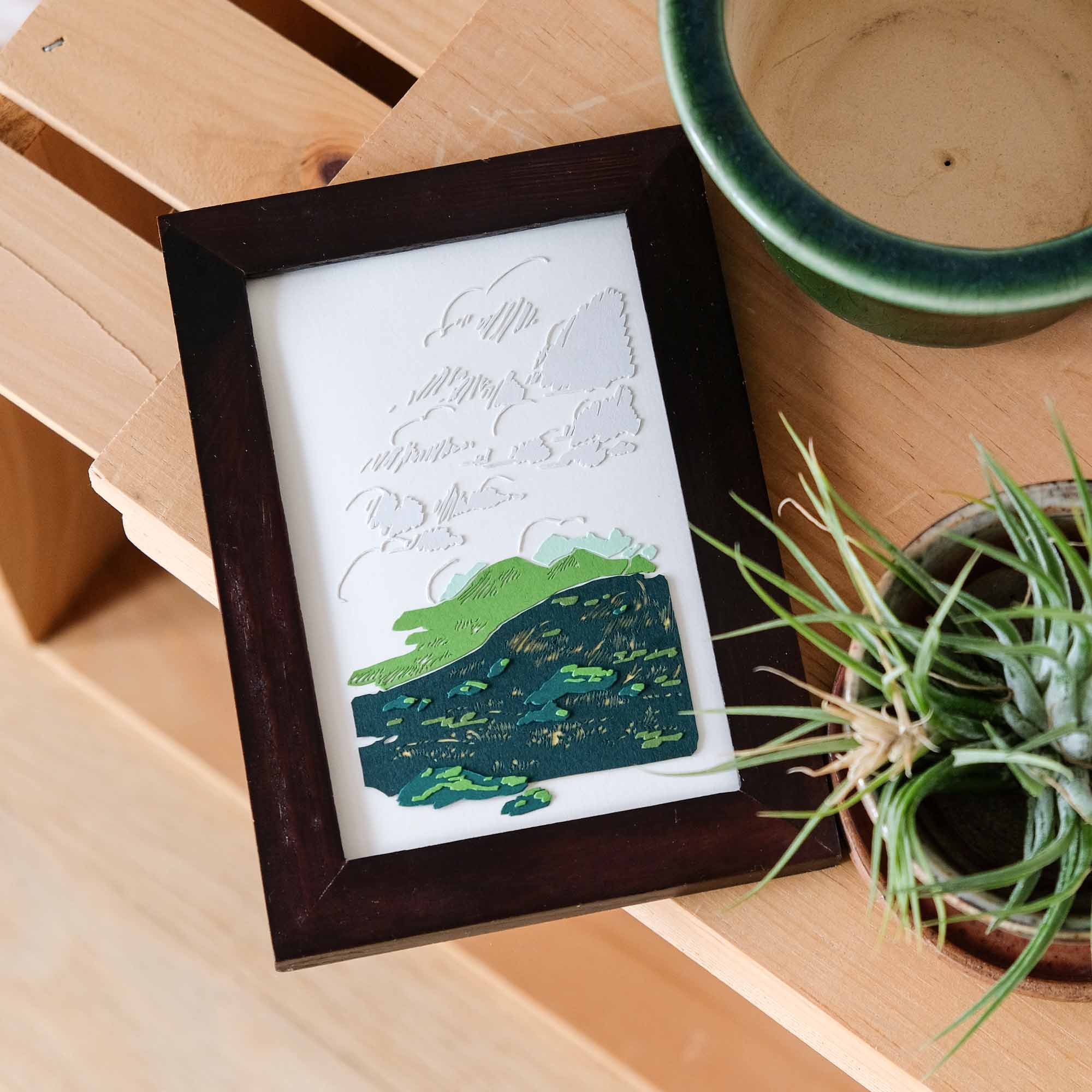 A paper scene depicts a dark green hill, covered with clusters of brush and fine strands of golden grasses, under gestural gray clouds. The artwork is closely framed with dark wood next to a small plant and layers of light wood.