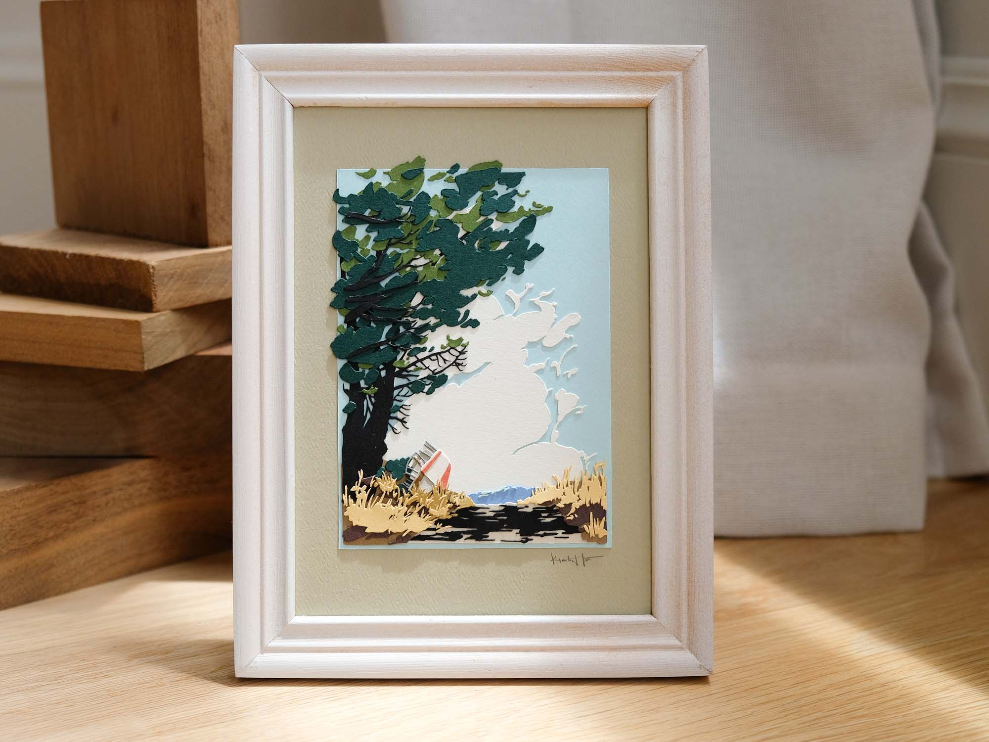 A framed cut paper piece depicts a tall green tree casting its dappled shade on dry grasses, which partially obscure a red and white boat beside a dirt road. The frame sits among wood decor and swatches of white fabric.