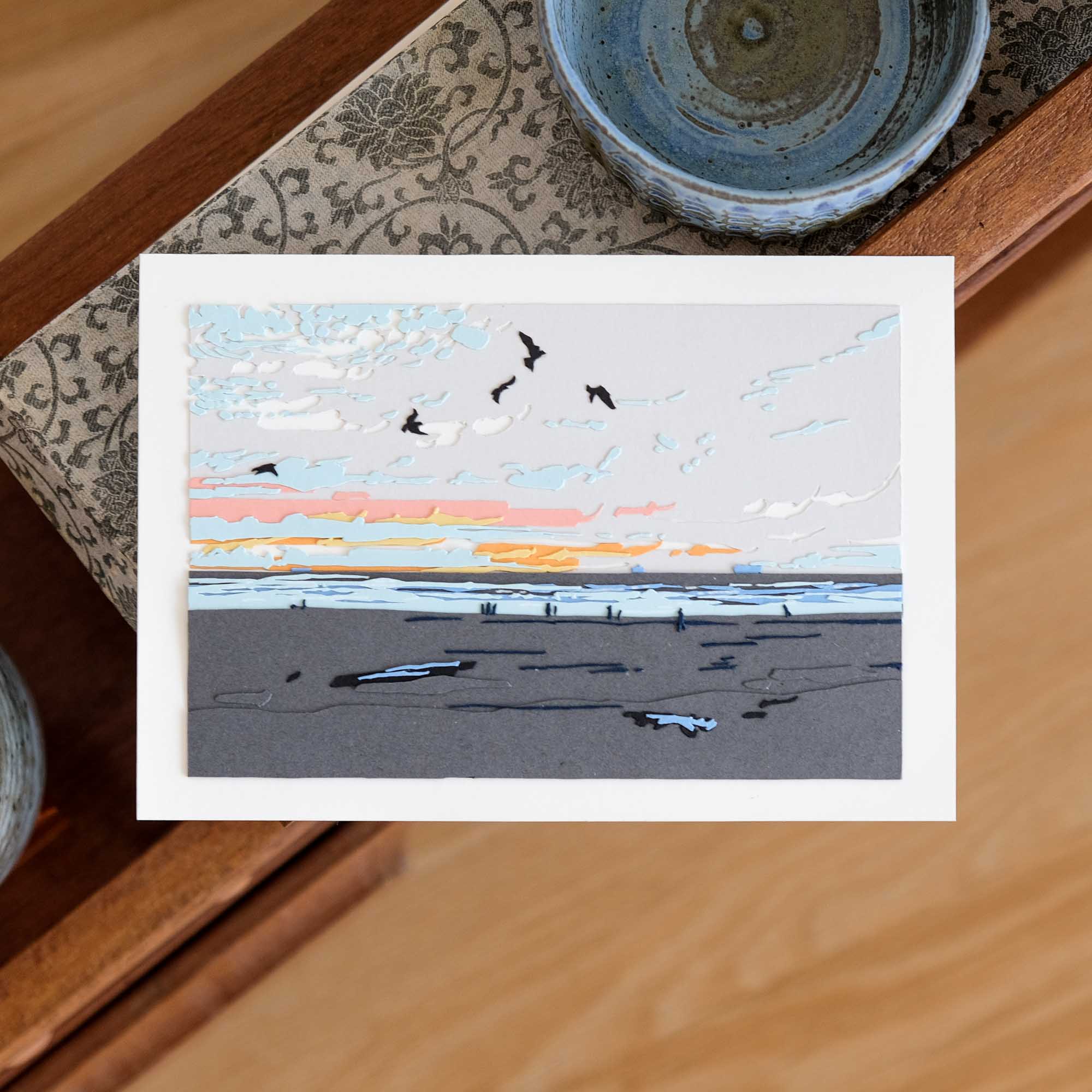 A beach of gray sand stretches towards a sliver of ocean underneath a pale sky. Paper birds are silhouetted against a paper sunset, while tiny paper humans cross the far side of the beach. The artwork lies on a wooden shelf with various ceramics and objects.