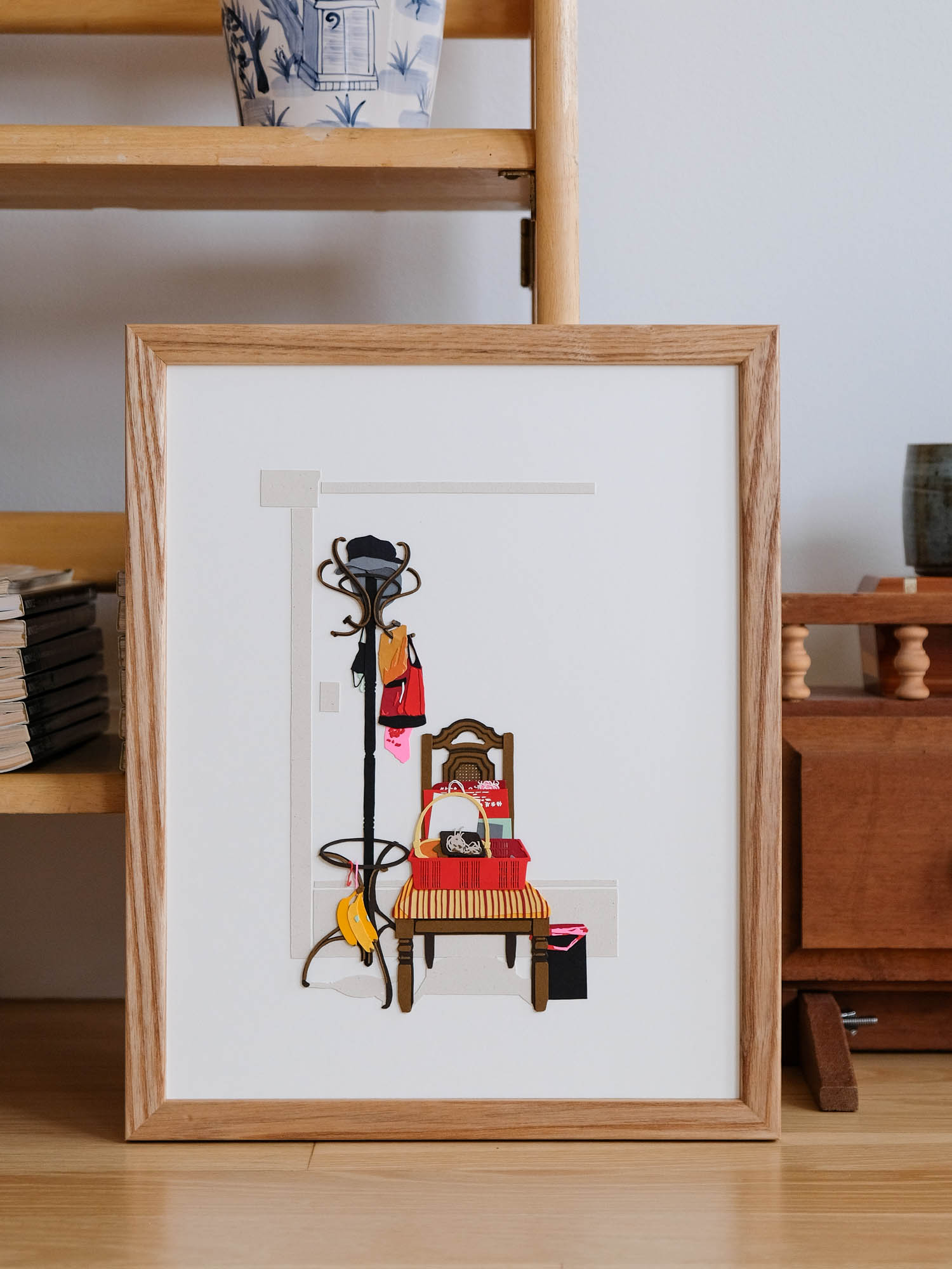 A paper coatrack stands next to a paper dining chair. Both are festooned with various bags, baskets, paper goods, and a clutch of bananas. Faint shapes hint at the architecture of the room beyond.